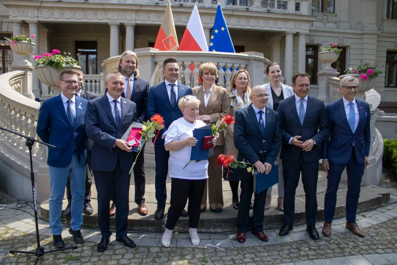 Grupa osób w garniturach i strojach formalnych stoi przed budynkiem z flagami.