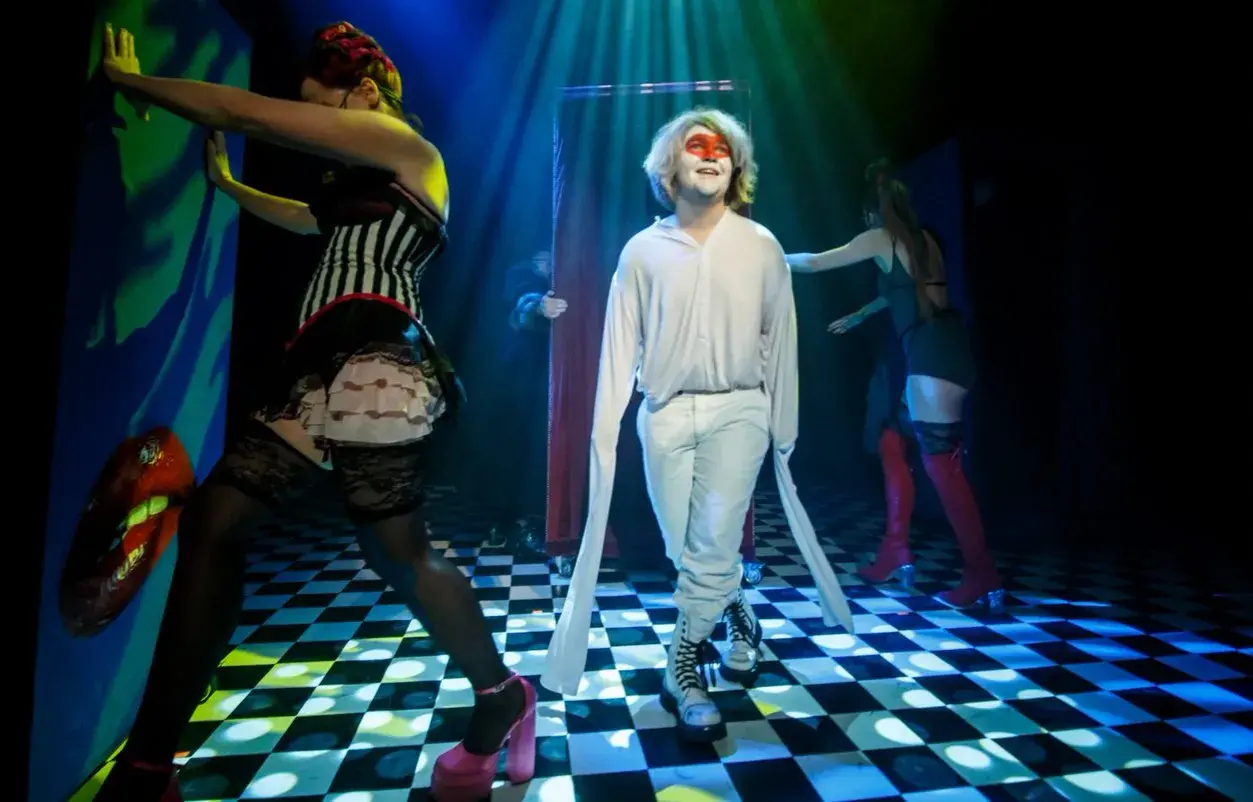Scena teatralna, gdzie młody mężczyzna w białym stroju i makijażu twarzy stoi pośród postaci w kostiumach, tworząc atmosferę tragifarsy.