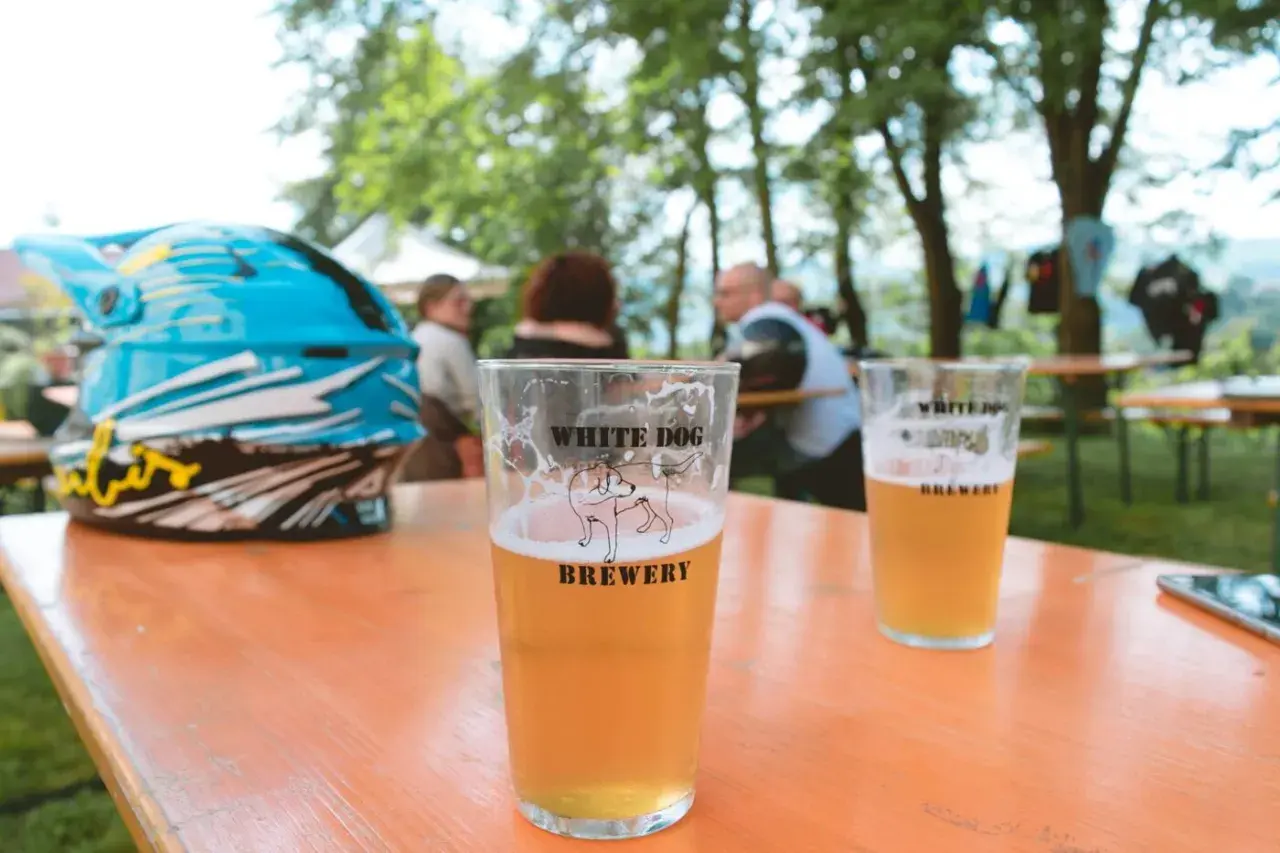 Bicchiere di birra con logo White Dog Brewery, accanto a un casco da moto blu, su un tavolo all'aperto.