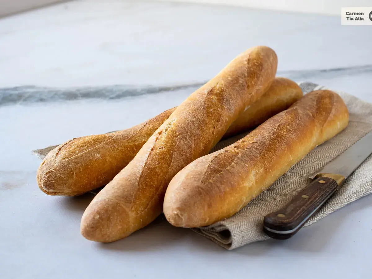 Tres baguettes doradas recién horneadas, listas para aprender como hacer baguette. Un cuchillo de madera descansa a su lado.