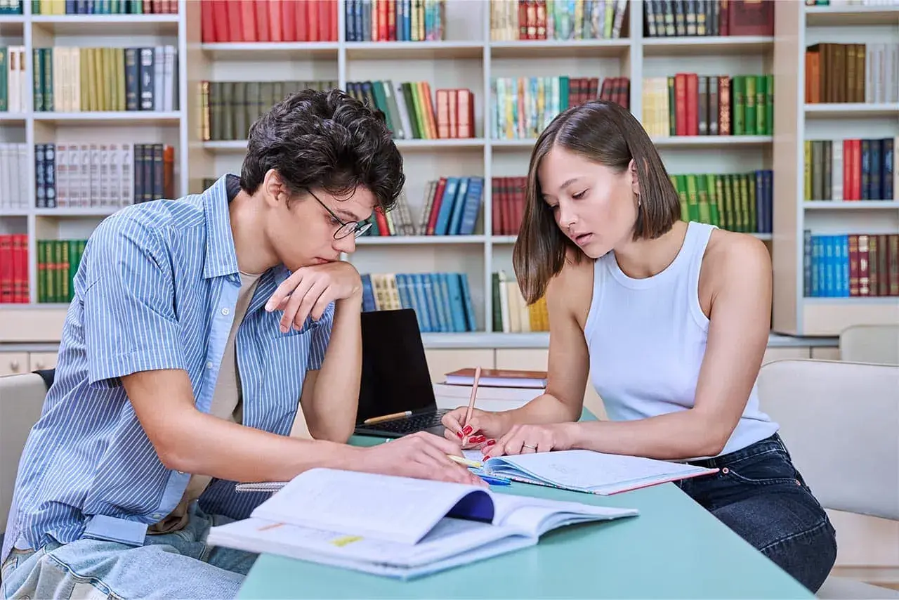 Dos personas jóvenes estudian juntas en una biblioteca, rodeadas de estanterías llenas de libros.