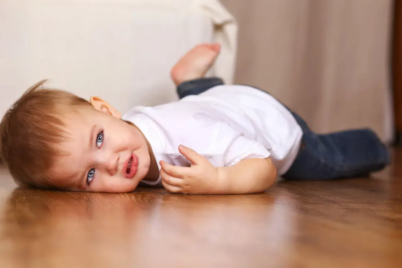Un bebé de 16 meses con ojos azules y cabello castaño yace en el suelo de madera, vestido con una camiseta blanca y jeans.