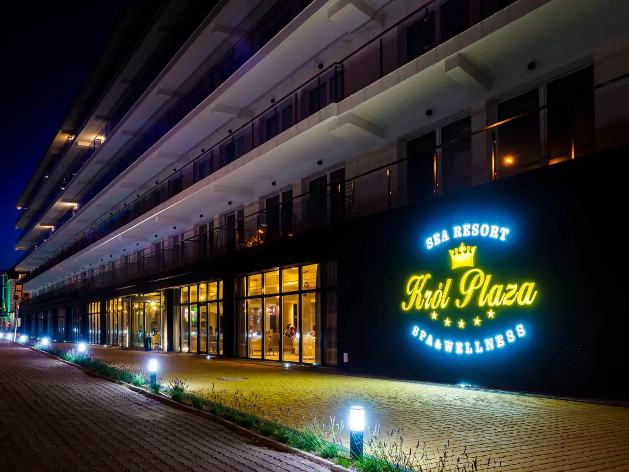 Nocny widok na nowoczesny hotel Król Plaza Spa & Wellness w Jarosławcu. Neonowy napis zachęca do sprawdzenia opinii o tym miejscu.