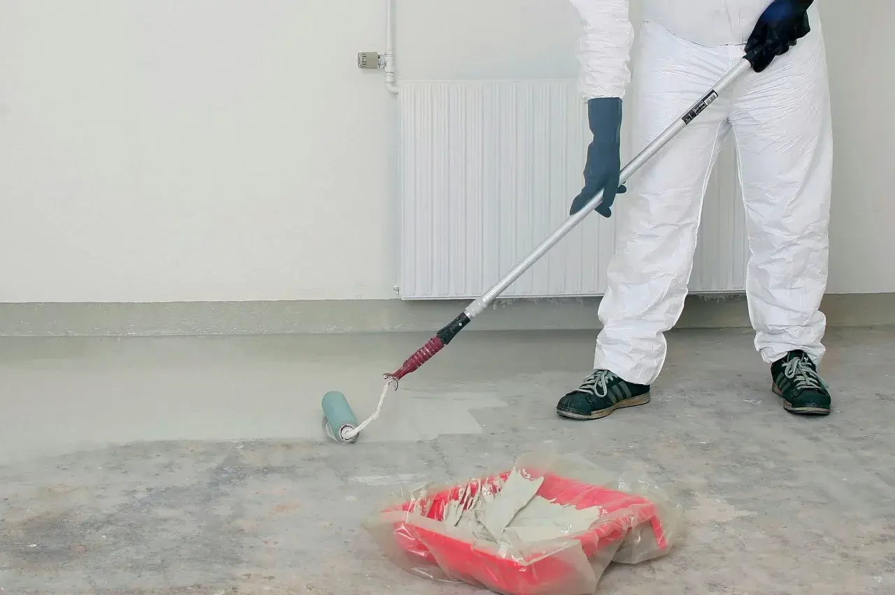 Person i skyddsdräkt målar betonggolv med roller. En färgtråg med ljus färg står på golvet.