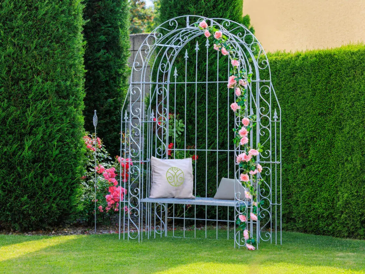 Ein weißer Rosenbogen mit rosa Rosen, Sitzbank und Kissen. Tolle Rosenbogen Gestaltungsideen für Ihren Garten.