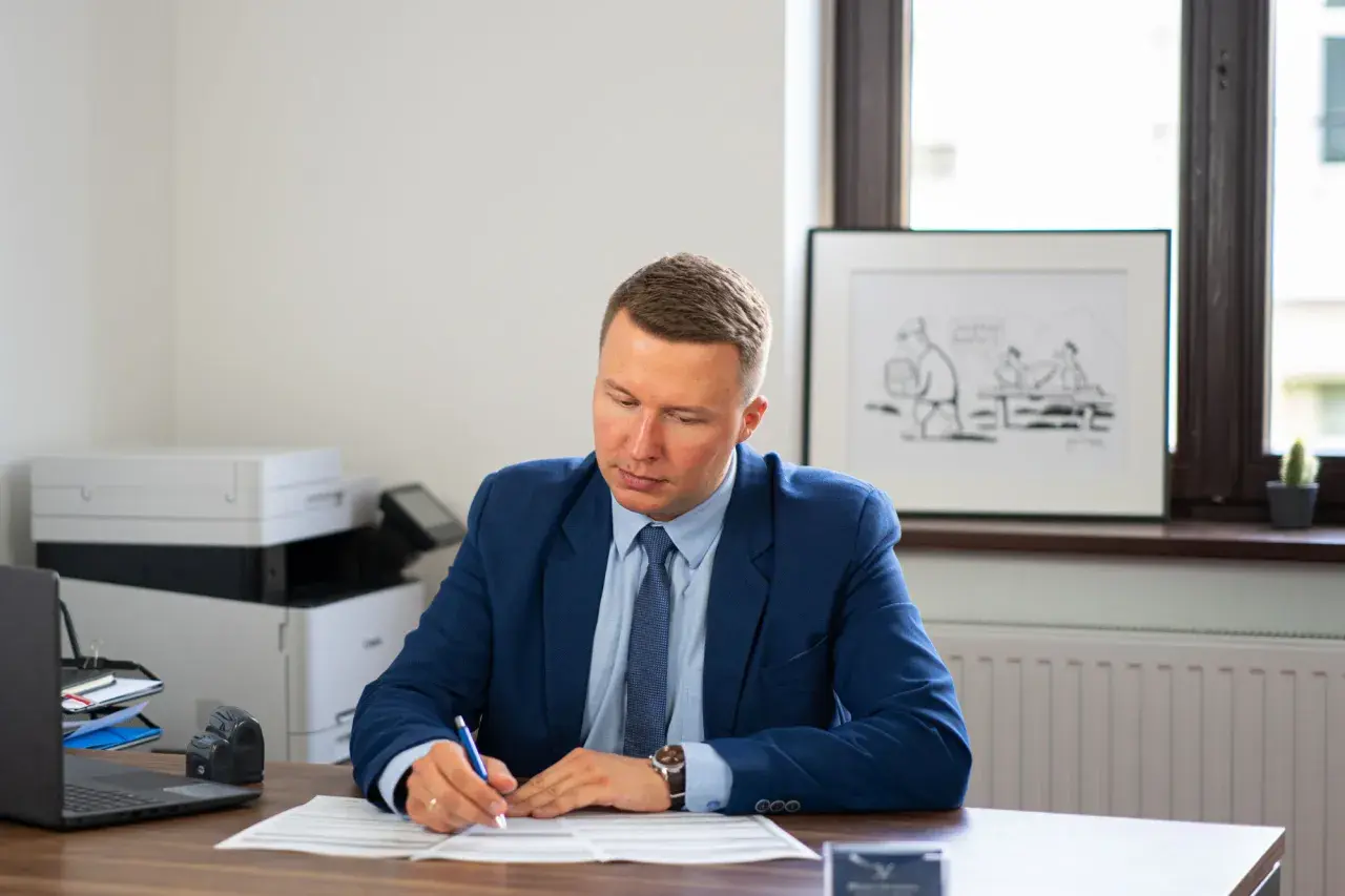 Mężczyzna w garniturze siedzi przy biurku i pisze na dokumentach.