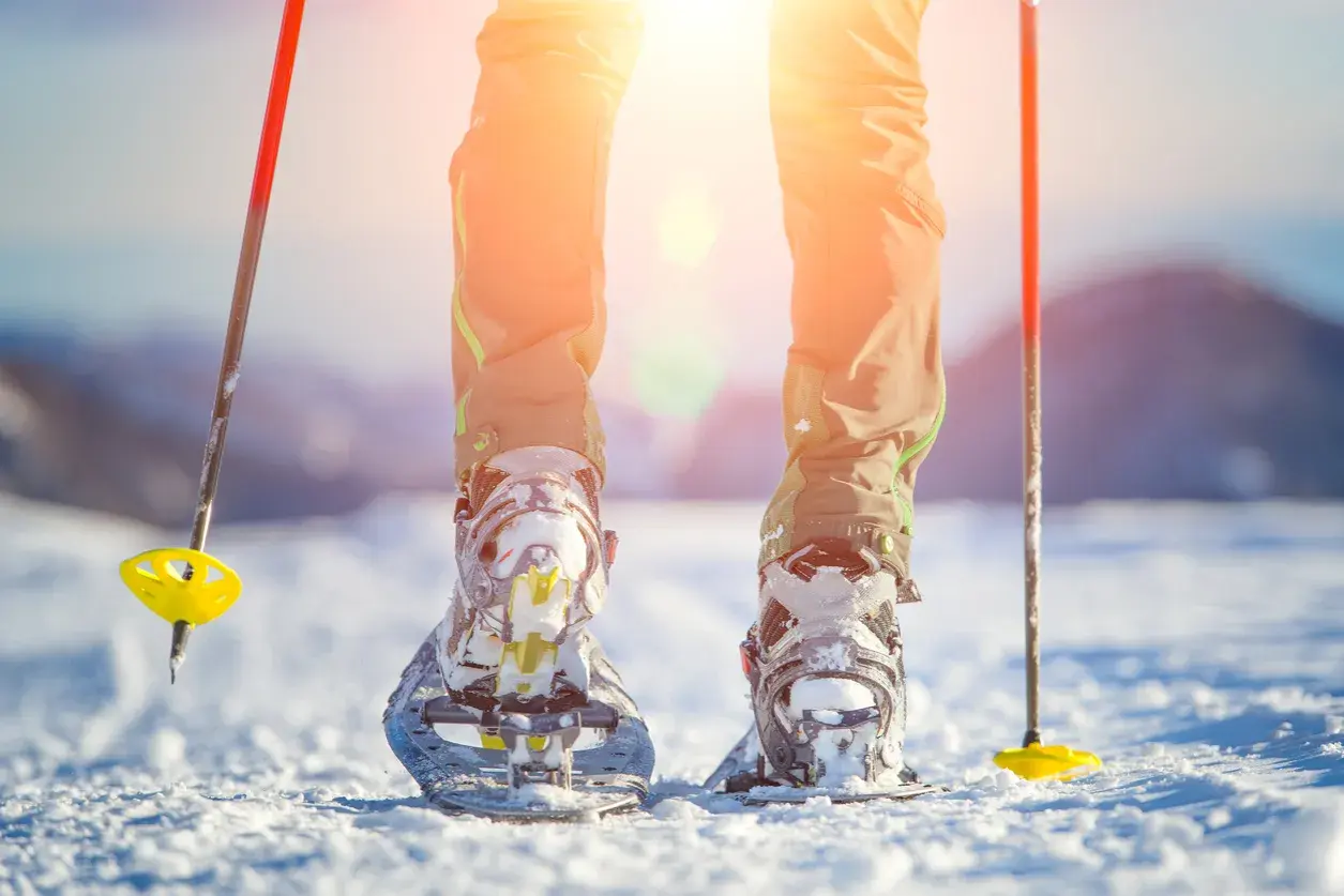 Piernas con raquetas de nieve y bastones listos para excursiones en la nieve bajo un sol brillante.