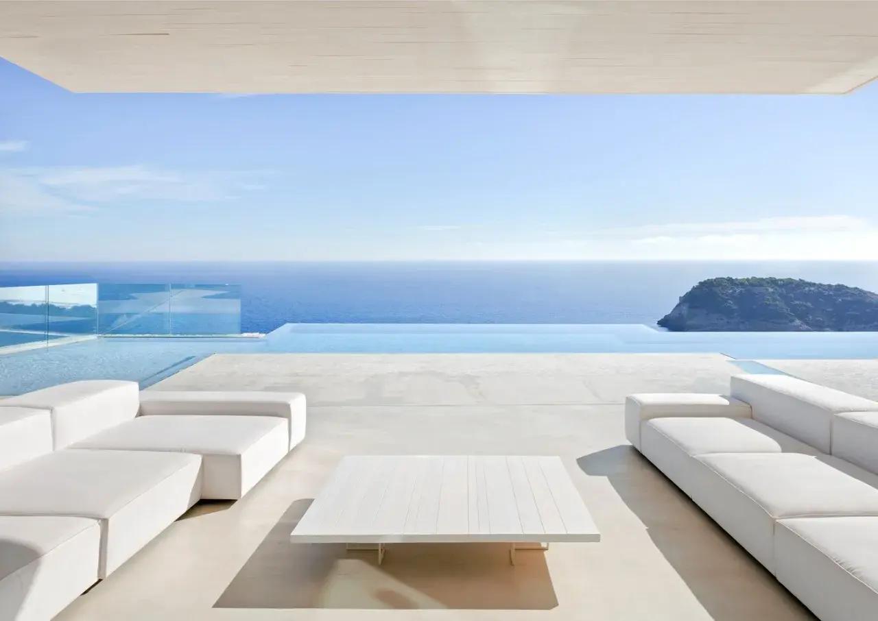 Terrasse moderne avec canapés blancs et table basse, bordée par une piscine à débordement et la mer bleue. Le **béton ciré autour de la piscine** crée une ambiance épurée et luxueuse.
