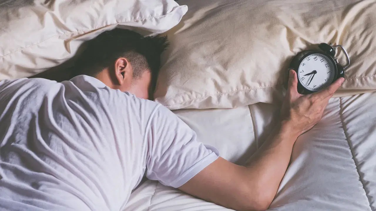 Mann schläft im Bett, Wecker liegt daneben. Wie viel Schlaf braucht man wirklich?