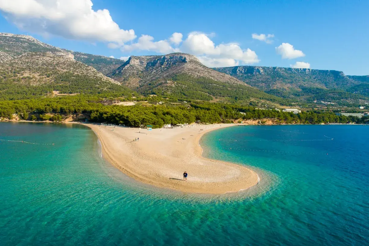 Una spiaggia a forma di lingua di sabbia dorata si estende nel mare turchese della costa croata, con montagne verdi sullo sfondo.