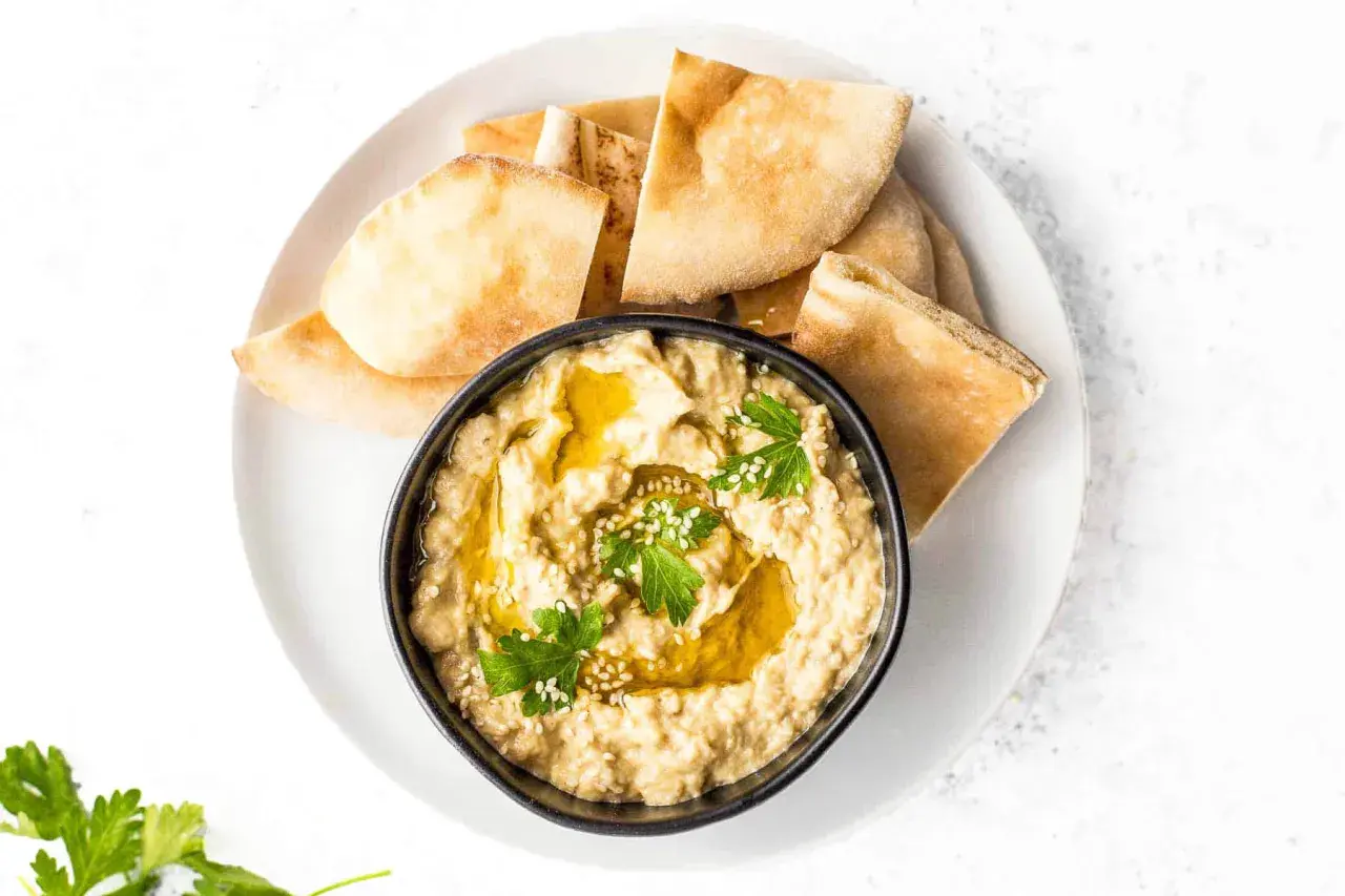Un bol de caviar d'aubergine crémeux, garni d'huile d'olive, de persil et de sésame, servi avec des morceaux de pain pita.