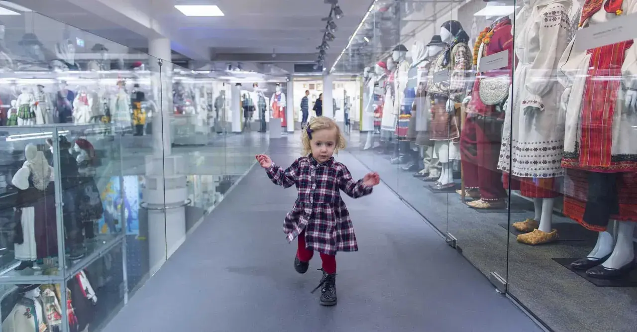 Mała dziewczynka biega po muzeum dla dzieci im. Janusza Korczaka, mijając gabloty z tradycyjnymi strojami.