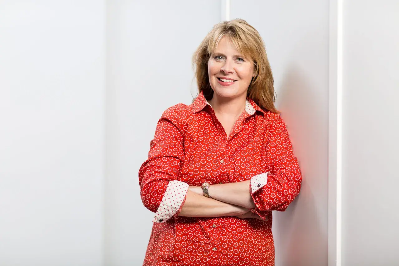 Petra Weber, eine lächelnde Frau mit blonden Haaren, trägt ein rotes Hemd mit Blumenmuster und hat die Arme verschränkt.