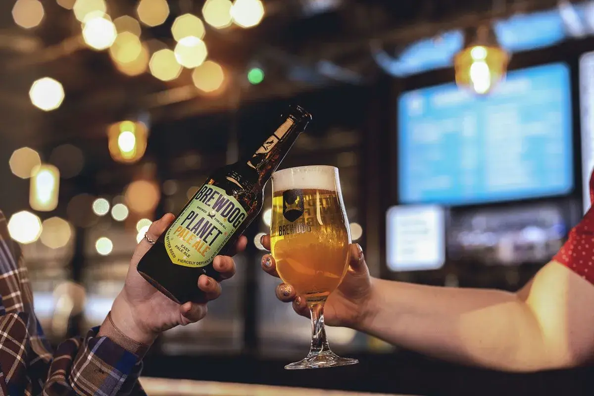 Brindisi con birra BrewDog Planet Pale Ale, perfetta per la Italy Beer Week. Luci soffuse e atmosfera da pub.