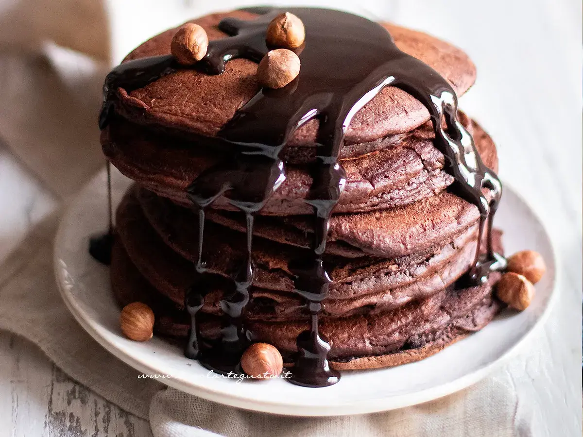 Pancake cacao golosi, impilati e ricoperti da una cascata di salsa al cioccolato fondente e nocciole intere.