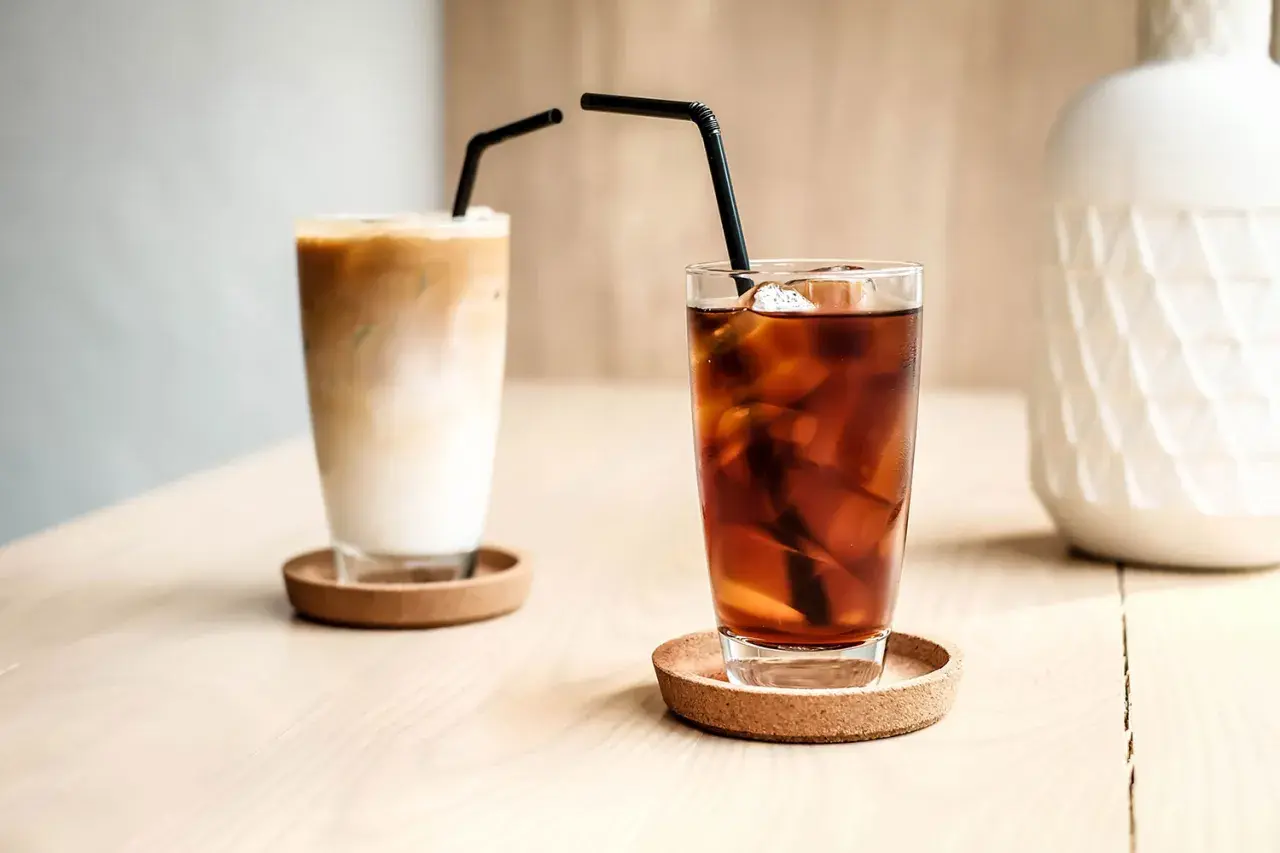 Zwei Gläser mit Eiswürfeln und Strohhalmen. Eines ist ein cremiger Eiskaffee, das andere ein dunkler Kaffee mit kaltem Wasser.