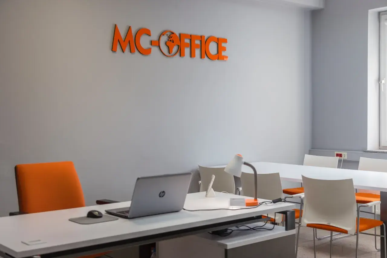 Pomarańczowe logo MC-OFFICE na szarej ścianie nad biurkiem z laptopem.