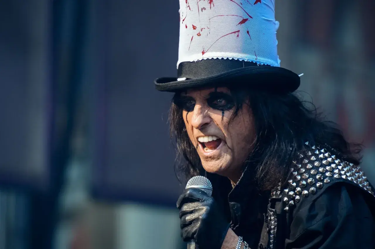 Alice Cooper w Polsce, w swoim charakterystycznym makijażu i z kapeluszem, śpiewa do mikrofonu.