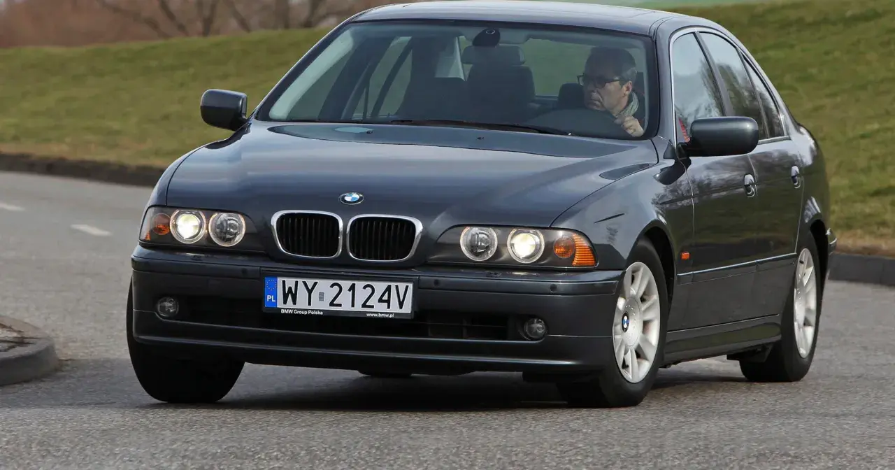 Ciemnoszary sedan BMW E39 z kierowcą w okularach jedzie po drodze.