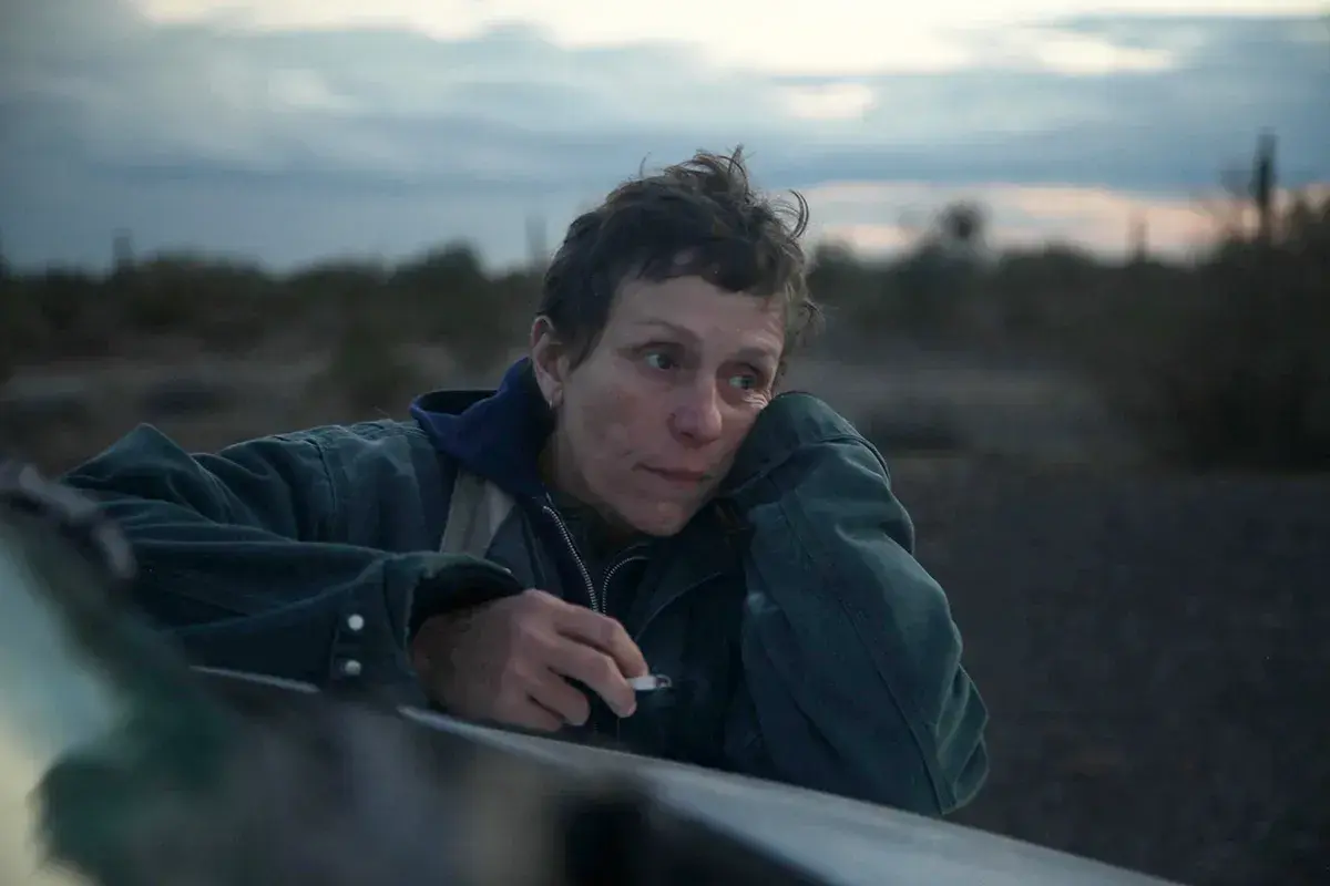 Skådespelare Frances McDormand lutar sig mot en bil i ökenlandskap, med en cigarett i handen.