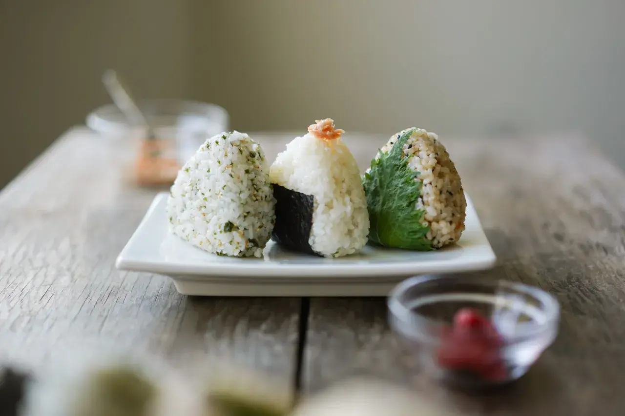 Three onigiri, made of rice, sit on a white plate. One is wrapped in nori, another topped with salmon, and the third with a green leaf.