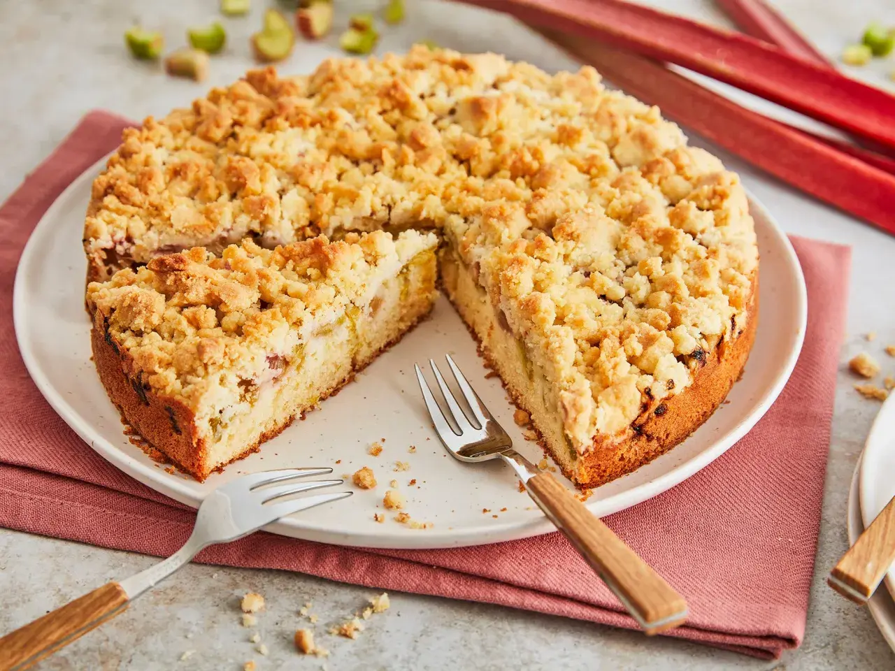 Ein saftiger Rhabarberkuchen mit Streuseln, bereit zum Genießen. Die roten Rhabarberstangen im Hintergrund versprechen Frische.