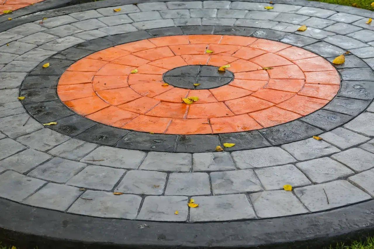 Kreisrunder Weg aus grau und orange gestrichenen Pflastersteinen, mit einigen Herbstblättern darauf.
