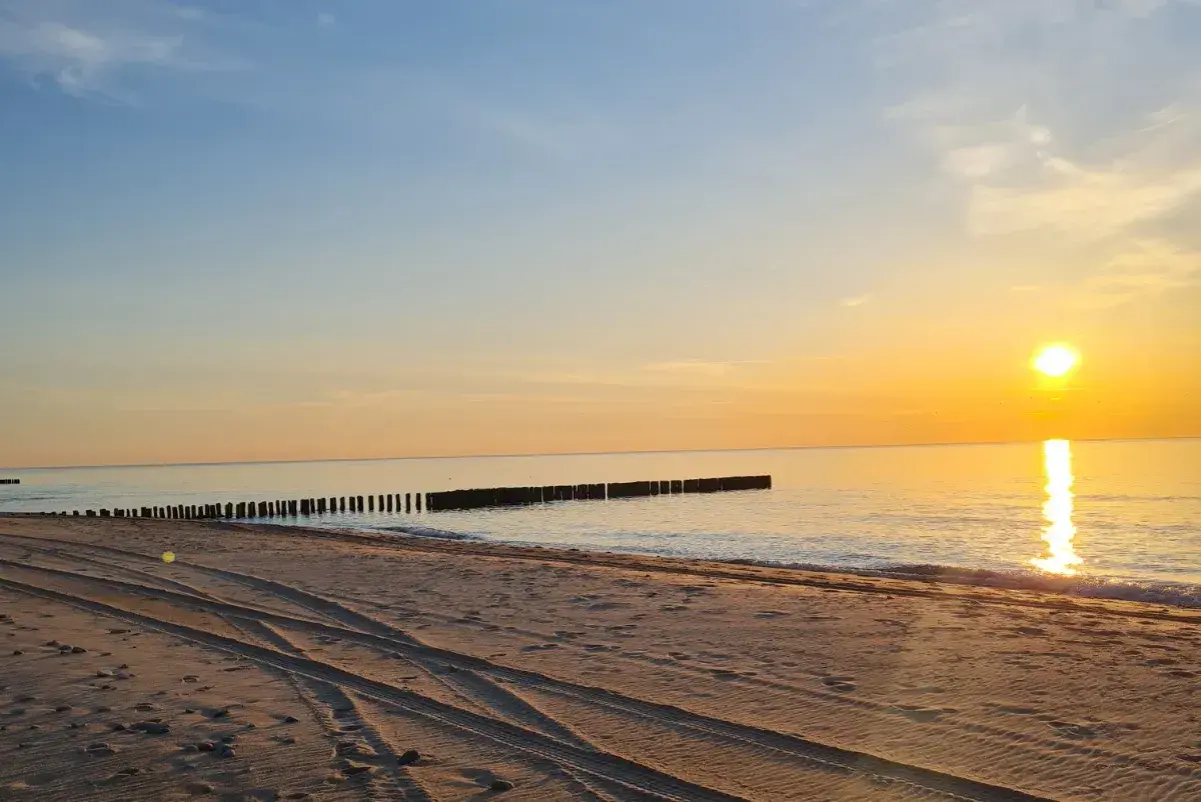 Zachód słońca nad morzem, piaszczysta plaża z widocznymi śladami opon, falochrony. Idealne miejsce na odpoczynek po podróży, nawet jeśli wicie dojazd PKP.