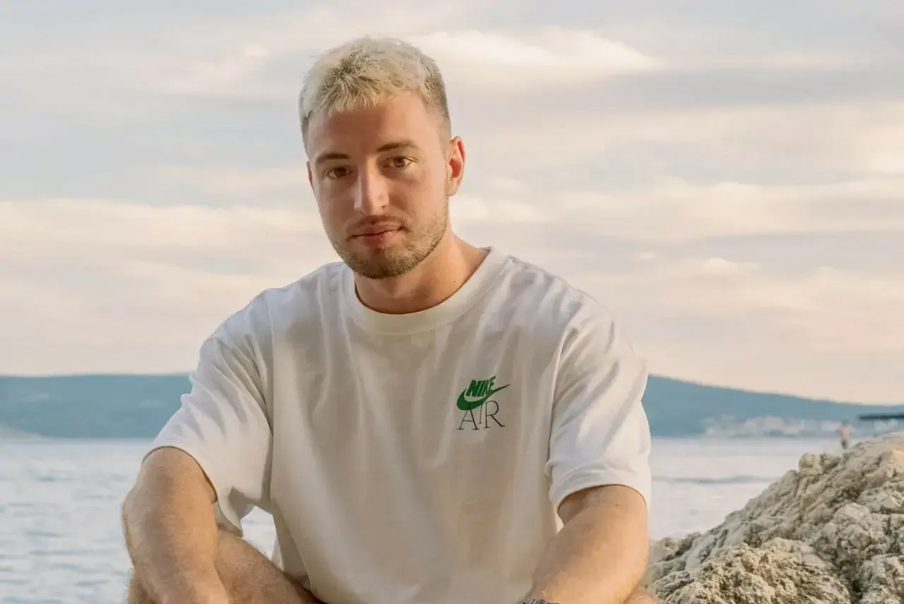 Edzio rap, z blond włosami i w białej koszulce, siedzi na plaży.