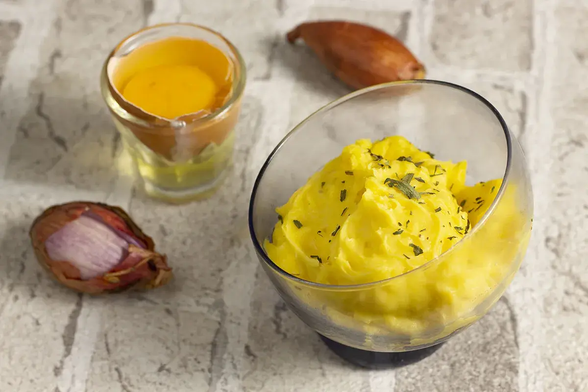 Una ciotola di salsa bernese cremosa, guarnita con erbe aromatiche, accanto a un tuorlo d'uovo e scalogni.