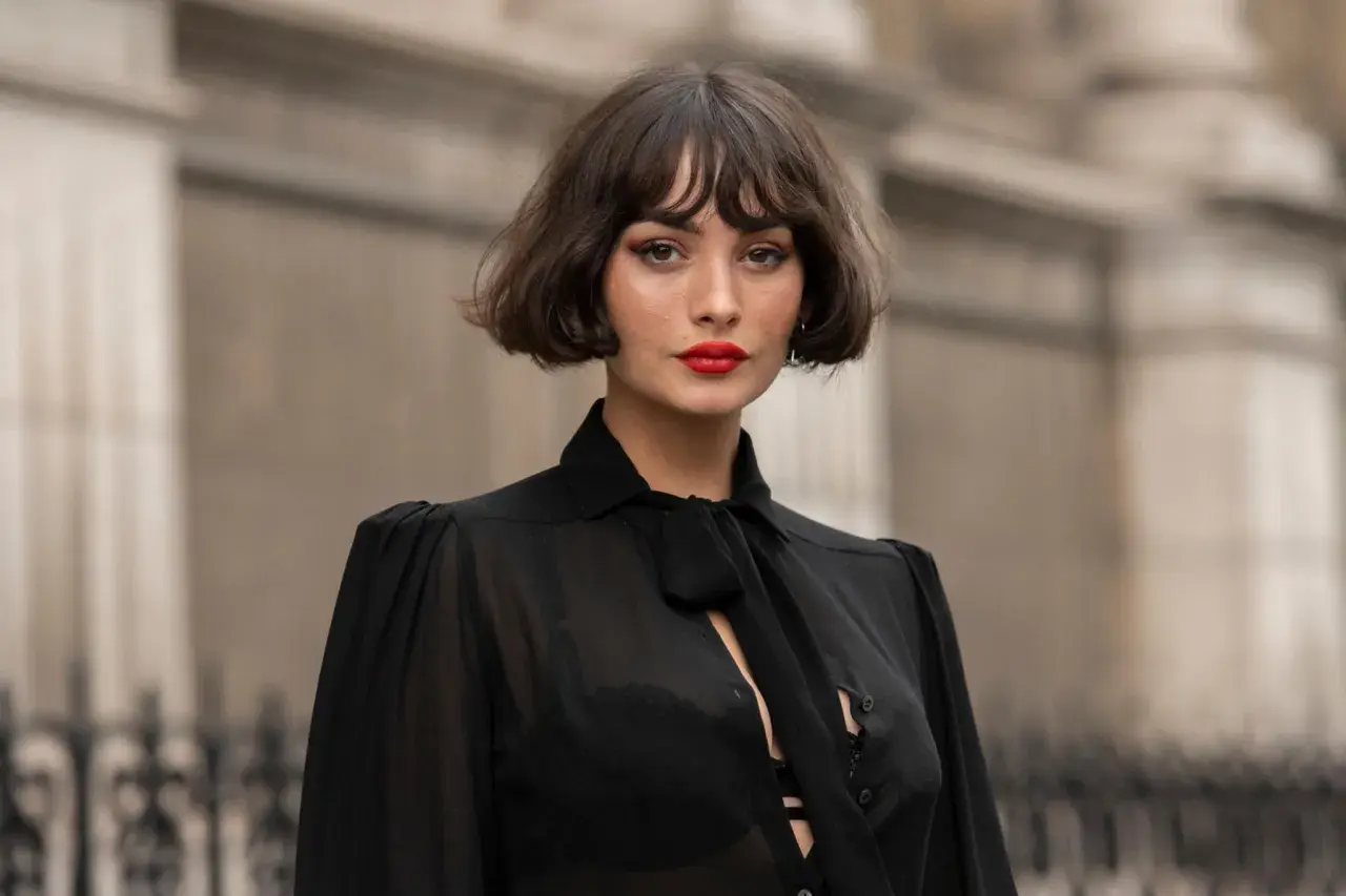 Eine Frau mit einem lässigen, **choppy bob** und rotem Lippenstift trägt eine transparente schwarze Bluse.