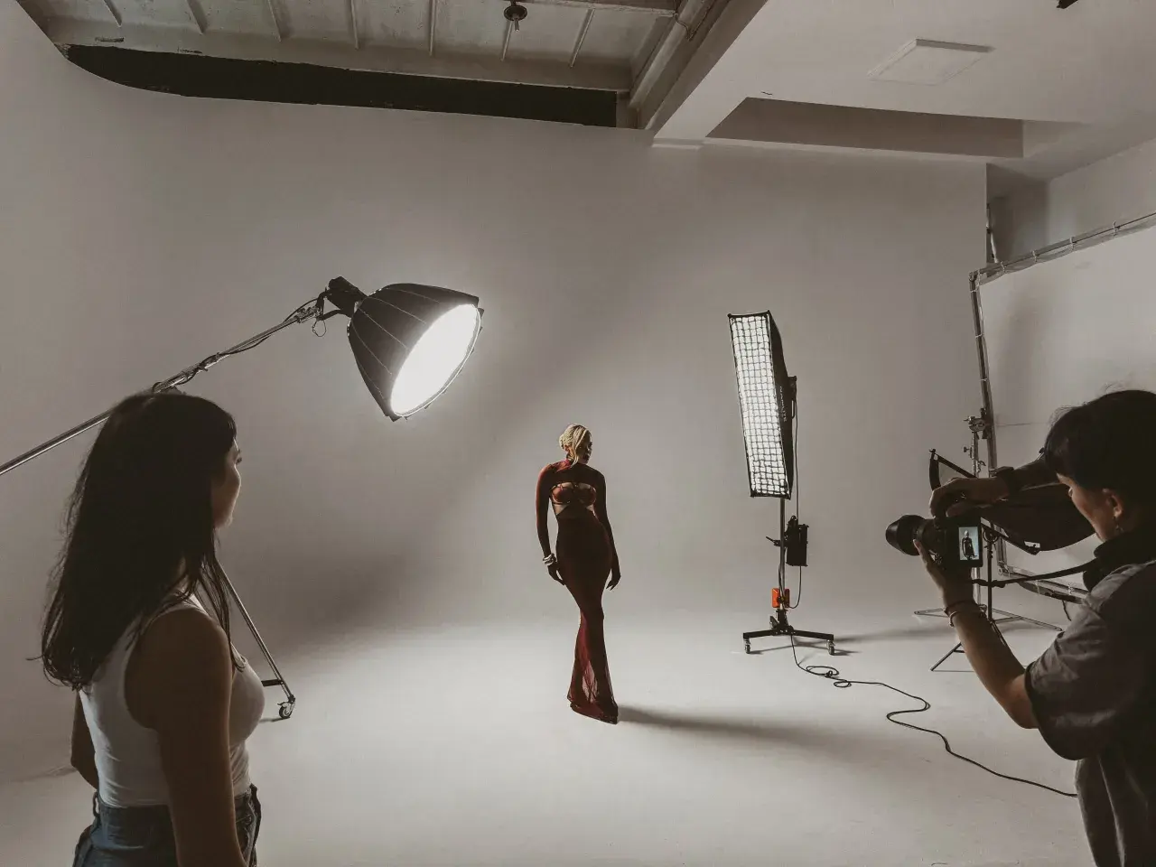 Modelka w czerwonej sukni pozuje na planie zdjęciowym. Fotograf z aparatem i asystentka przygotowują się do sesji.