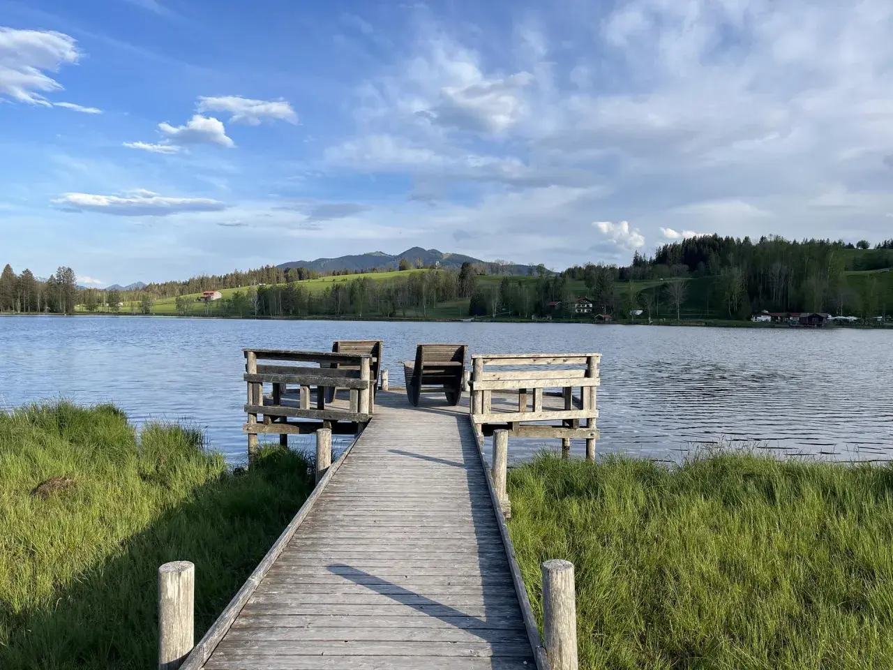 Holzsteg mit Sitzgelegenheiten am bayerischen See, umgeben von grünen Wiesen und bewaldeten Hügeln unter blauem Himmel.