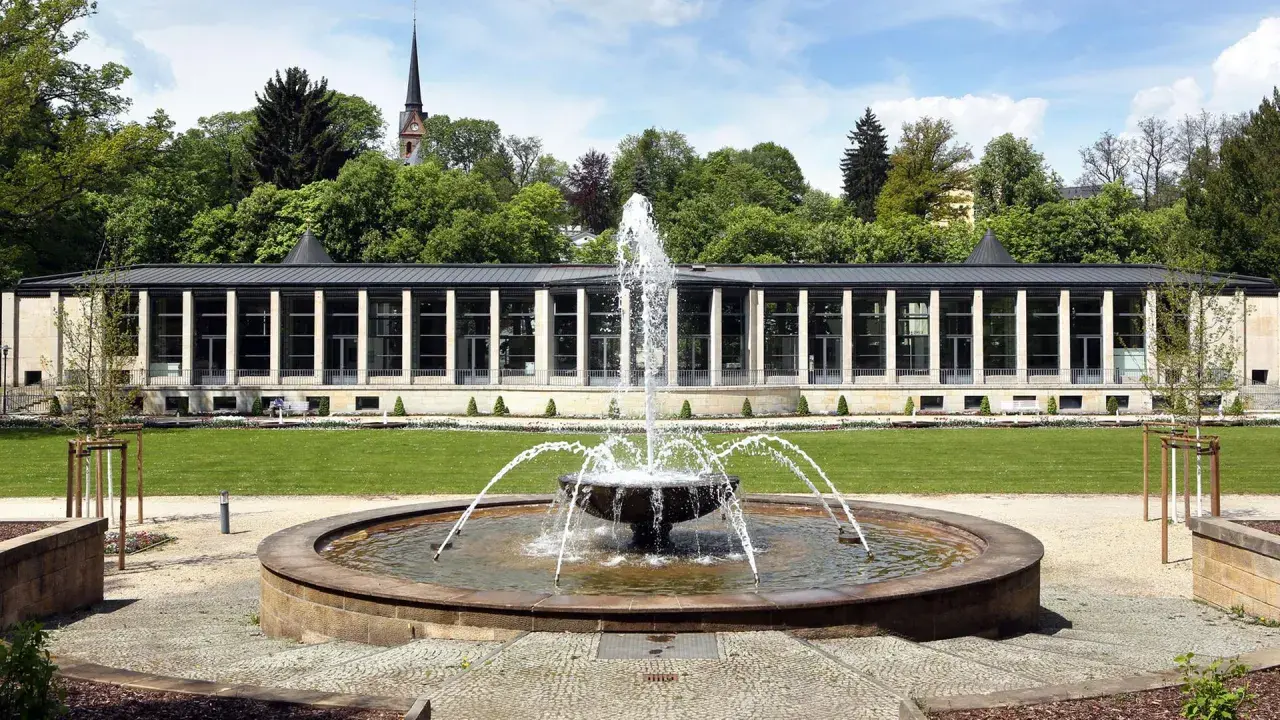 Ein Brunnen sprudelt vor einem modernen Gebäude mit Säulen und Glasfronten. Im Hintergrund ein Kirchturm und Bäume. Ein idyllischer tschechischer Kurort.