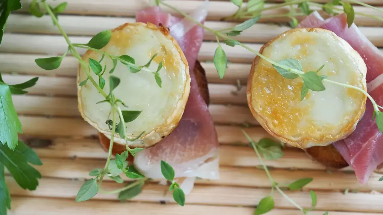 Dos bocaditos con crujiente de queso de cabra fundido y jamón serrano, adornados con hierbas frescas.