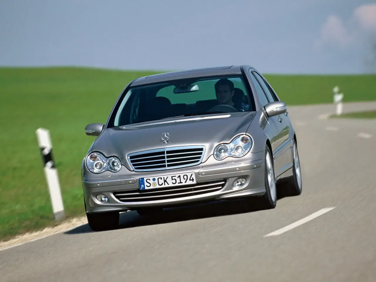 Srebrny Mercedes W203 1.8 Kompressor z kierowcą za kierownicą jedzie po krętej drodze wśród zielonych wzgórz.
