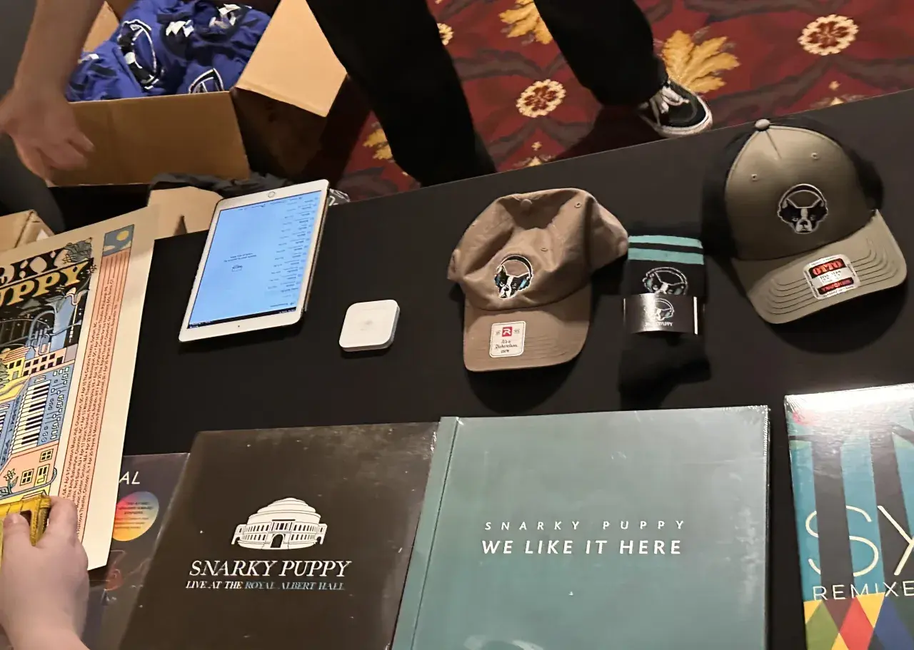 Snarky Puppy merchandise: vinyl records, hats, socks, and a tablet displaying a setlist.