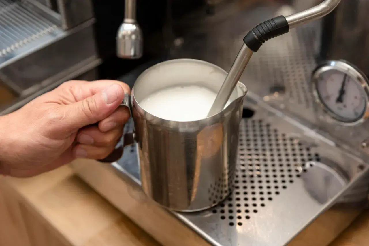 Ręka trzyma metalowy dzbanek z mlekiem, które jest spieniane przez dyszę ekspresu. Tak przygotowane mleko jest kluczowe, czy do cappuccino dodaje się mleko.