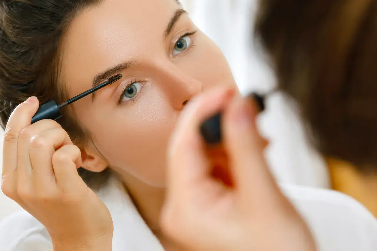 Une femme applique du mascara sur ses sourcils, préparant son maquillage bagage cabine pour un voyage.
