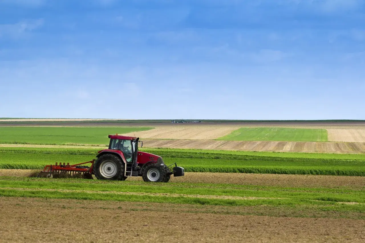 Tracteur rouge travaillant dans un champ, préparant le sol pour le semis direct. Paysage agricole sous un ciel bleu.
