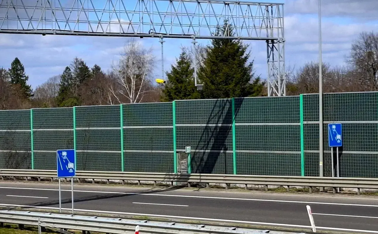 Znak informuje o odcinkowym pomiarze prędkości na A1. Za zieloną barierą dźwiękochłonną widać drzewa i niebo.