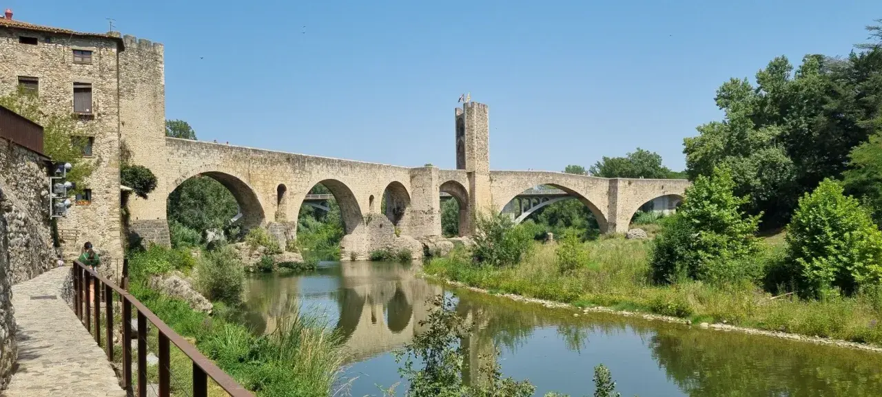 Puente medieval de Besalú, parte de una ruta histórica. Reflejo en el río y vegetación exuberante.