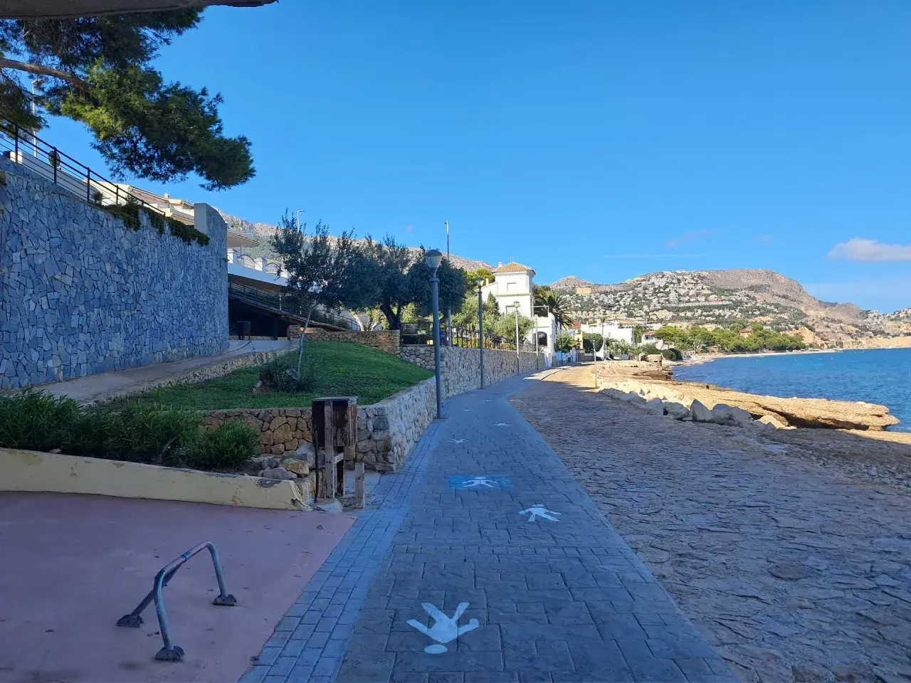 Paseo marítimo con siluetas de personas en la cala del Soio. El mar azul y las montañas de fondo crean un paisaje idílico.