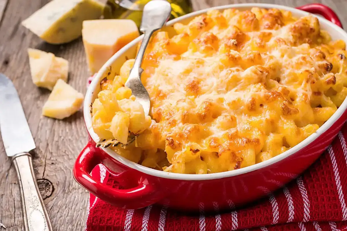Mac and cheese dorato e filante in una teglia rossa, con pezzi di formaggio e una forchetta pronta a servirlo.