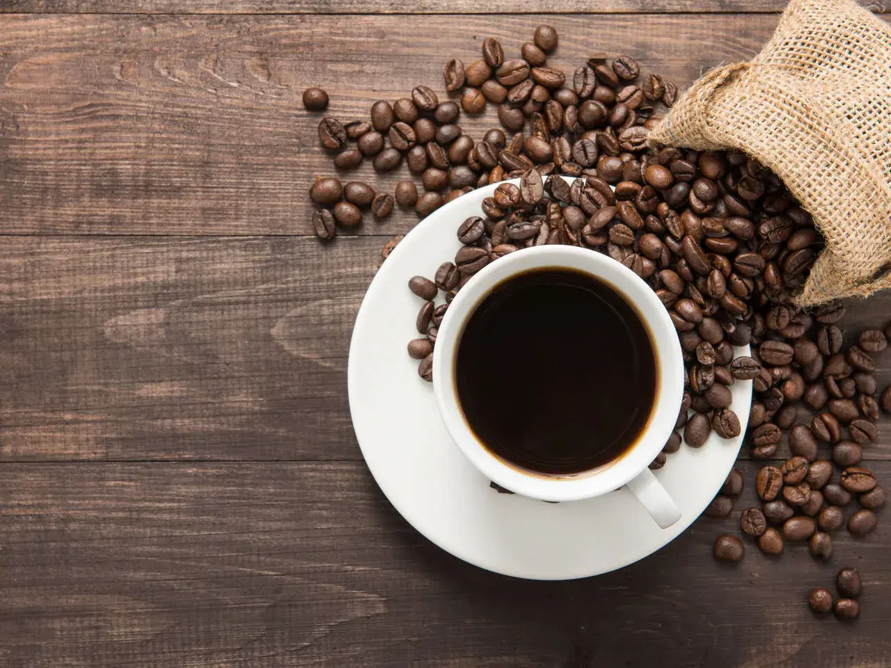 Aromatic coffee beans spill from a burlap sack onto a wooden table, next to a white cup filled with dark coffee. Poczuj kofeina działanie!