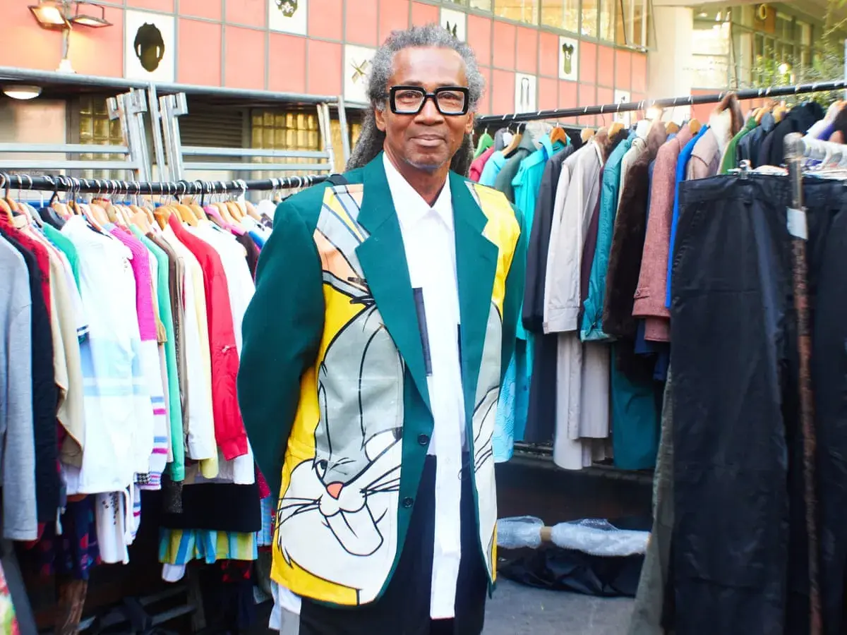 Man in a Bugs Bunny jacket stands by racks of colorful clothes, a testament to creative clothes upcycling.