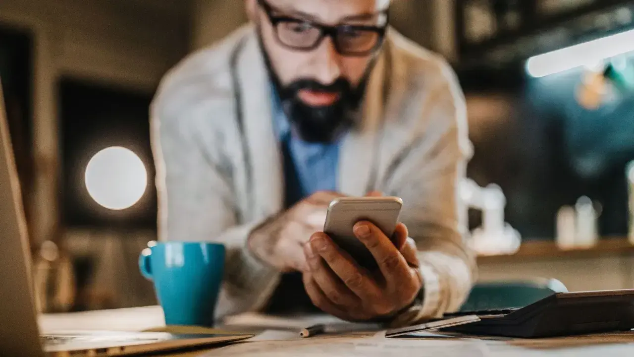 Mann mit Brille und Bart tippt auf Smartphone. Neben ihm eine blaue Tasse und ein Taschenrechner. Er sucht vielleicht, was ist die bankleitzahl.