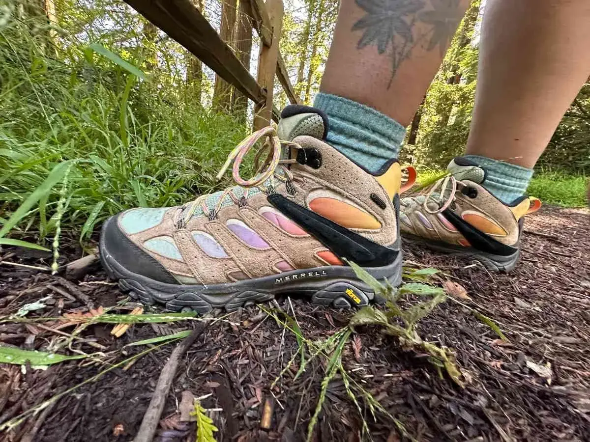 A person's feet wearing tan Merrell hiking boots with colorful accents and teal socks on a forest trail. These boots fit snugly, offering support for the hike.