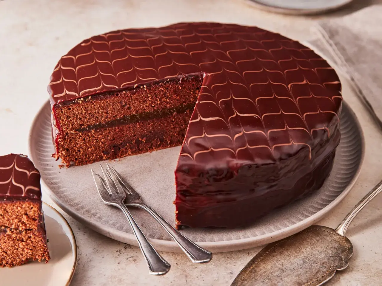 Eine glänzende Sacher Torte mit Schokoladenglasur und einem Stück Marmeladenfüllung. Gabeln liegen bereit.
