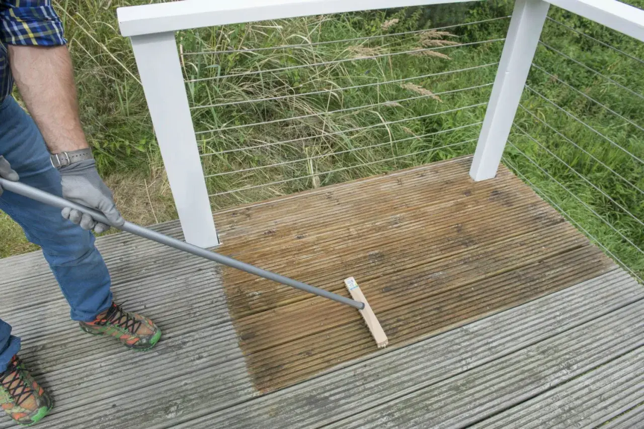 Mann reinigt mit einem Besen die Holzterrasse. Die eine Hälfte ist schon nass und dunkler, die andere noch trocken.
